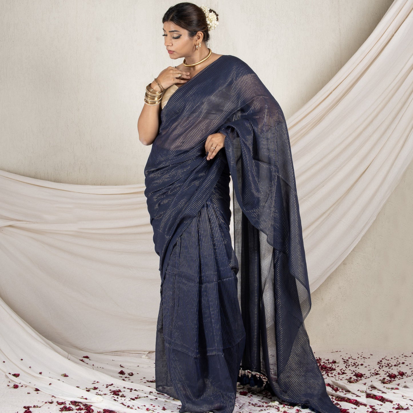 Woman in a dark blue saree standing against a white draped curtain with red flower petals on the floor.