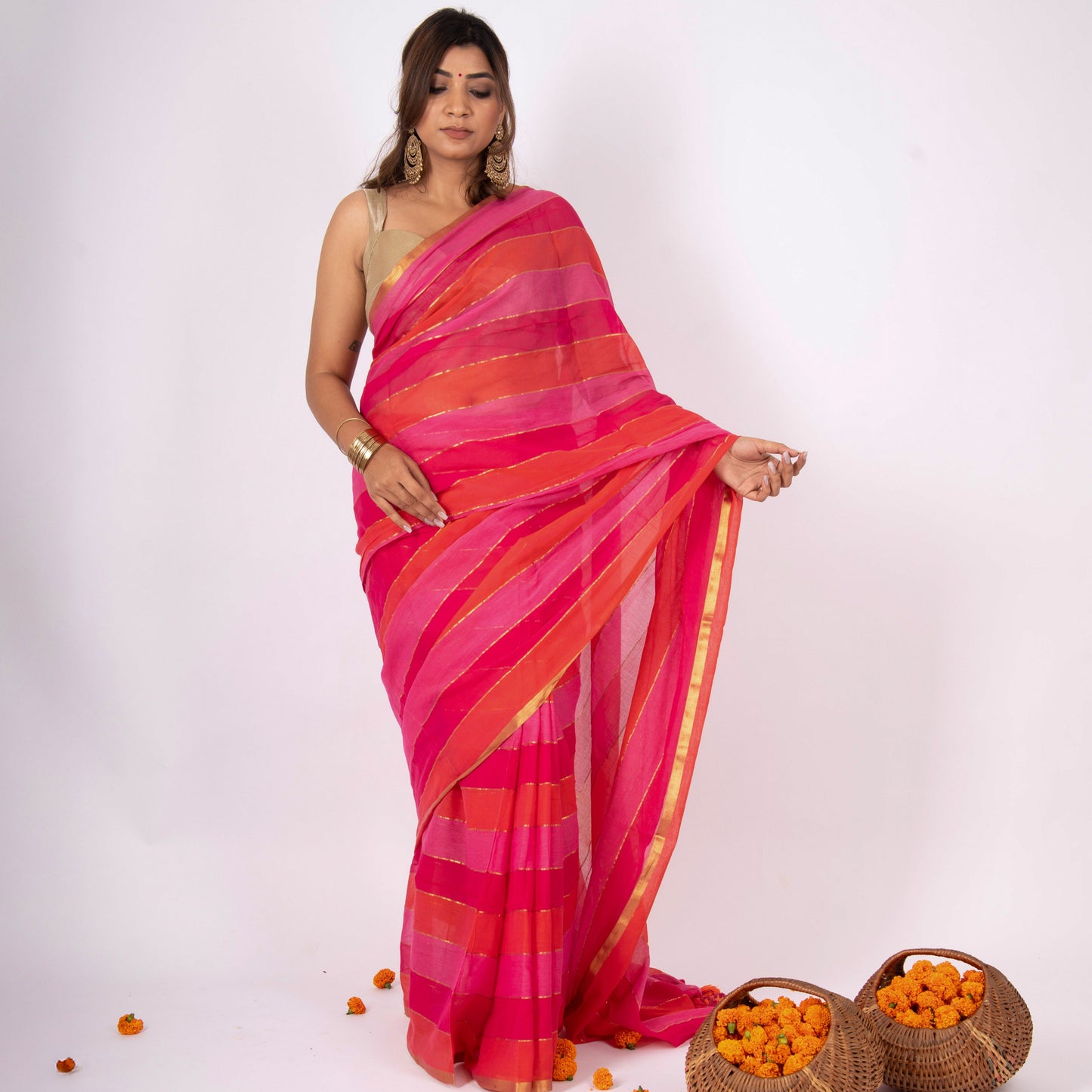Woman wearing a vibrant pink and orange saree with two baskets of oranges on a white background