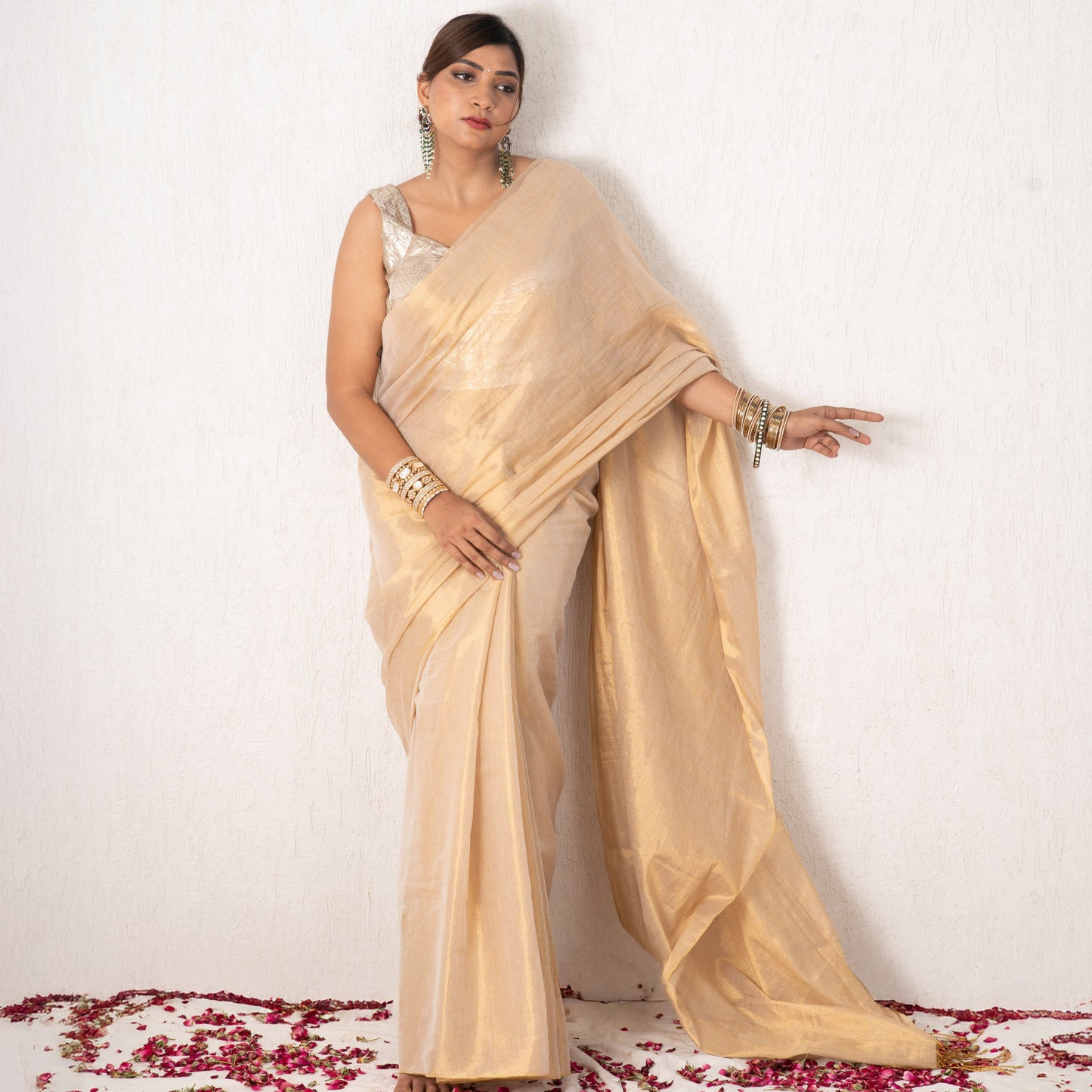 Woman in a beige saree standing on a white background with red flower petals at the base.
