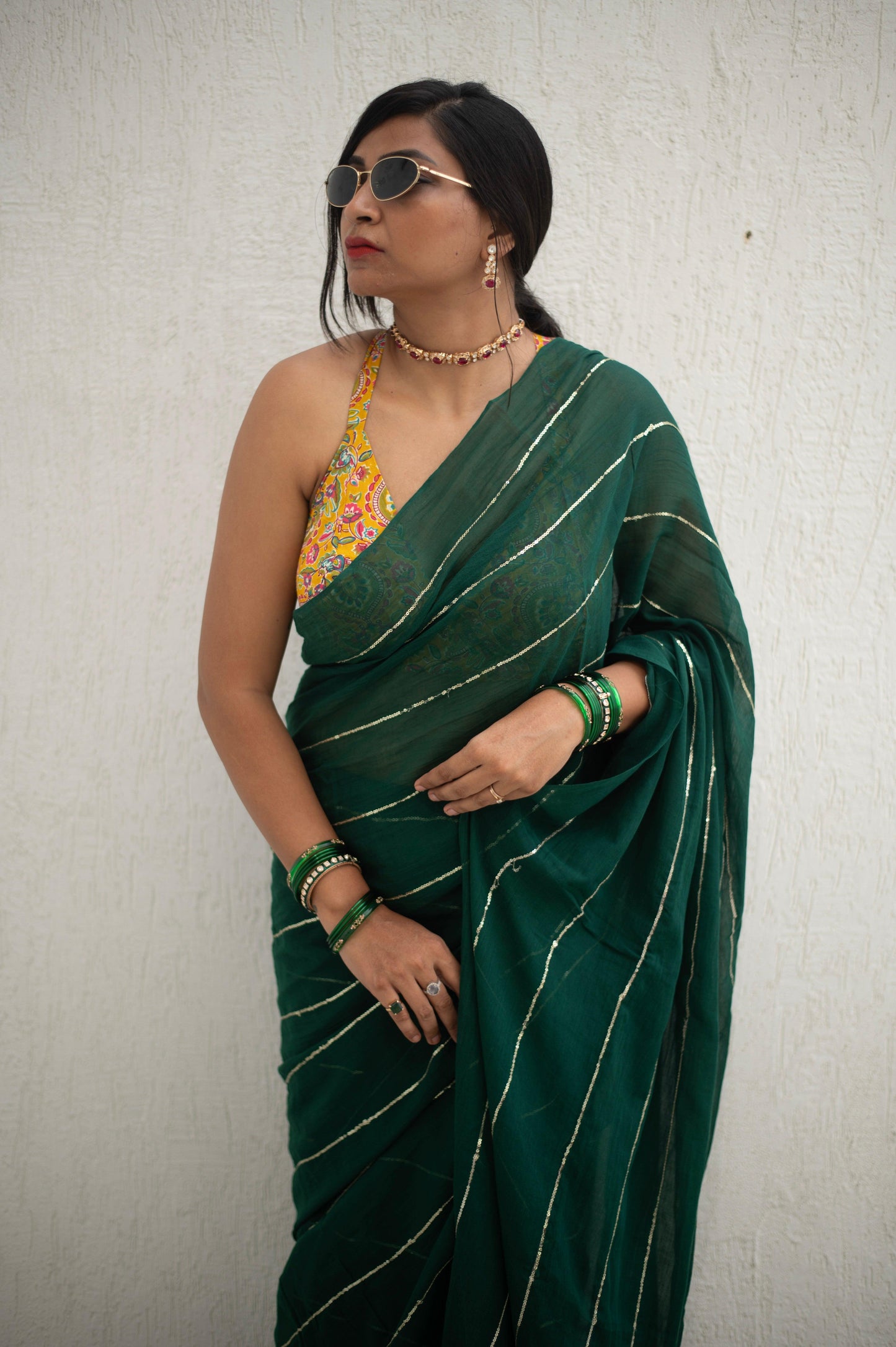 A woman modeling a green saree with sequins and a floral pattern, accessorized with sunglasses and a necklace.