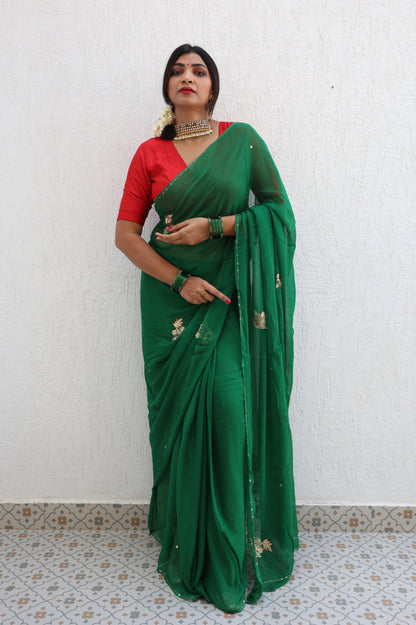 Woman wearing a green saree with a red blouse against a white wall.
