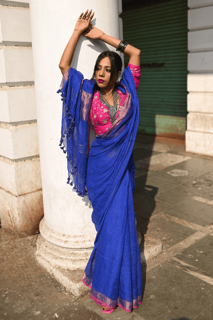 A woman showcasing a blue linen saree with pink border and intricate zari highlights, standing near a column.