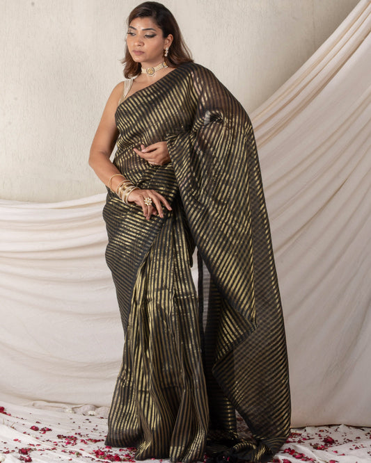 Woman in a black and gold saree standing against a white draped curtain with red flower petals on the floor.