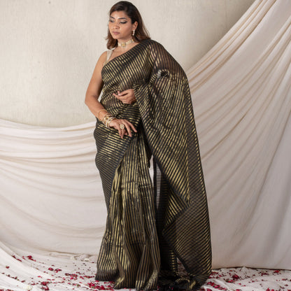 Woman in a black and gold saree standing against a white draped curtain with red flower petals on the floor.