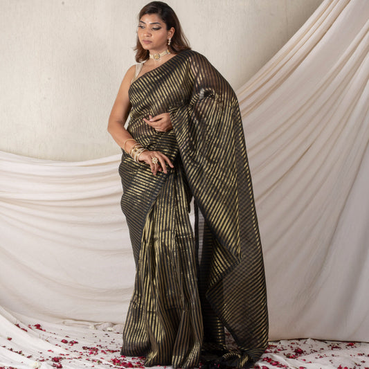 Woman in a black and gold saree standing against a white draped curtain with red flower petals on the floor.