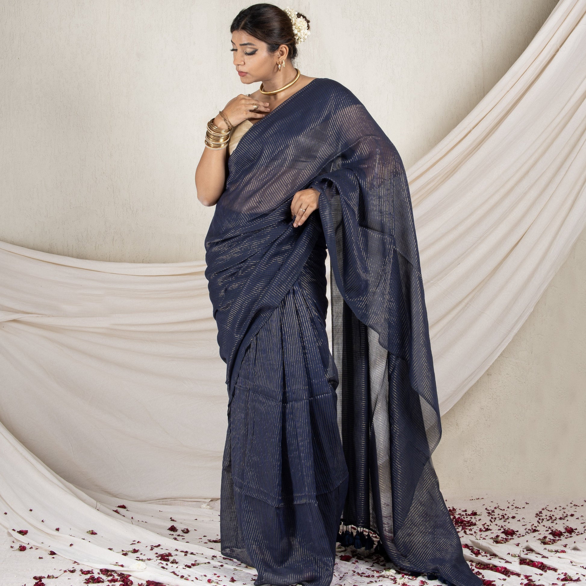 Woman in a dark blue saree standing against a white draped curtain with red flower petals on the floor.