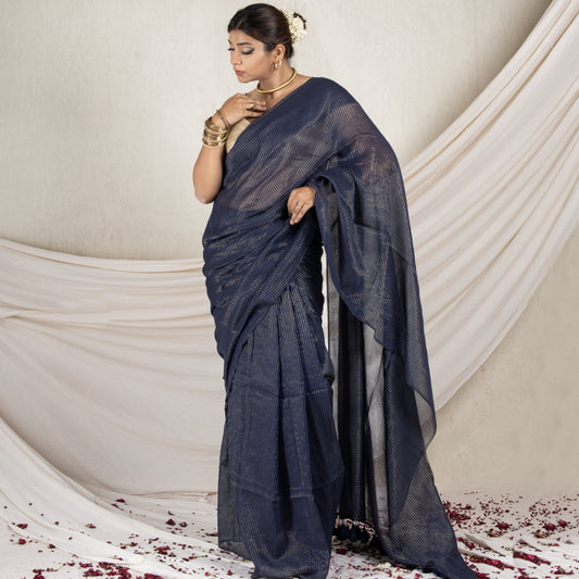Woman in a dark blue saree standing against a white draped curtain with red flower petals on the floor.