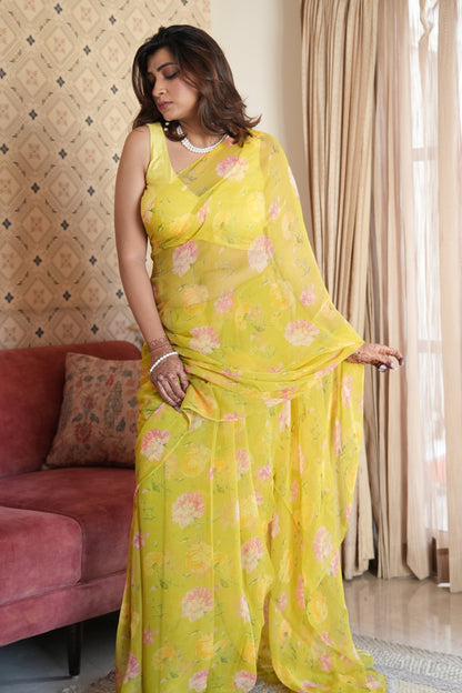 Woman in a yellow saree with floral patterns standing in a room with a pink sofa and patterned wallpaper.