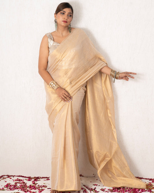 Woman in a beige saree standing on a white background with red flower petals at the base.