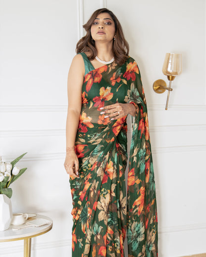Woman wearing a floral saree standing in a room with a white wall and a small table with flowers.