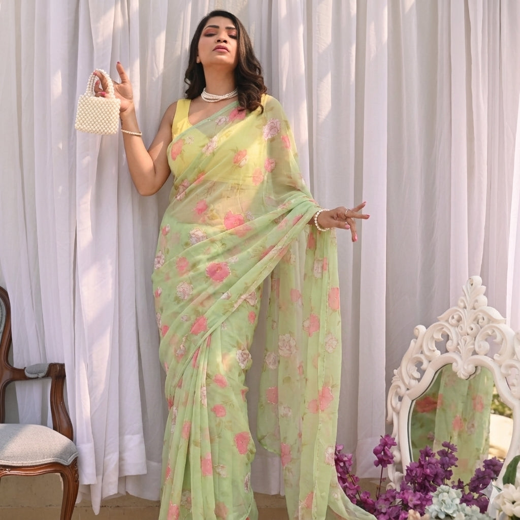 Woman in a light green floral saree standing in front of white curtains with a decorative mirror and flowers.