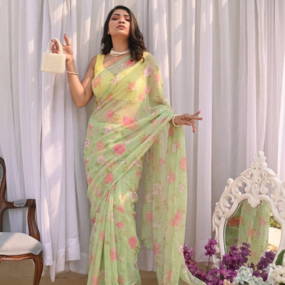 Woman in a light green floral saree standing in front of white curtains with a decorative mirror and flowers.