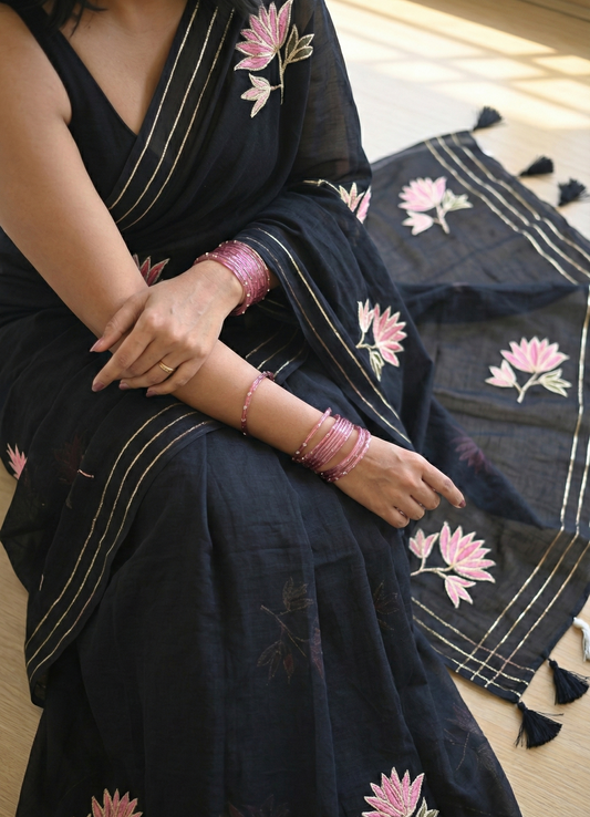 Person wearing a black saree with pink floral embroidery, sitting on a matching fabric background.