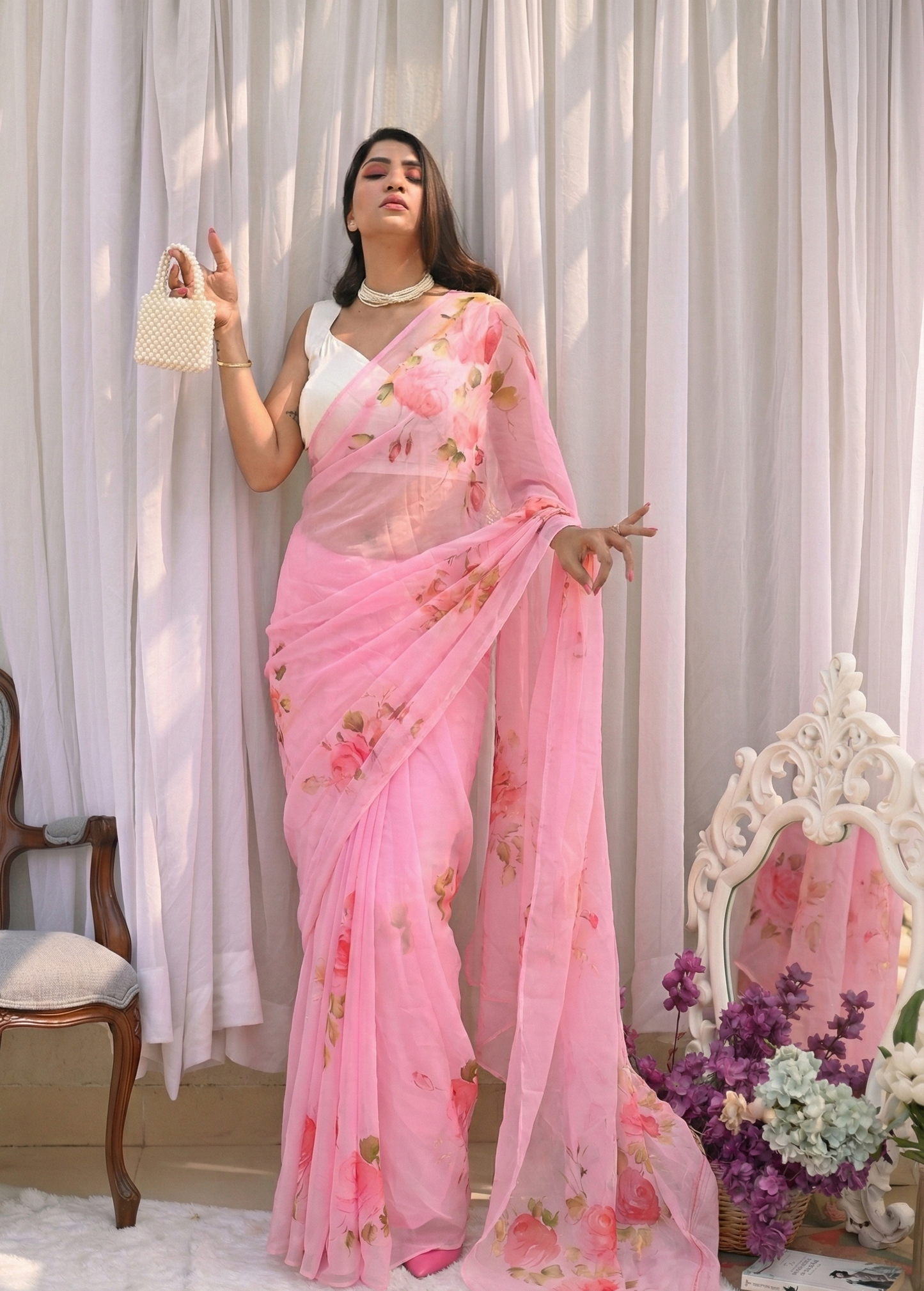 Woman wearing a pink floral saree with a white blouse against a white curtain background.