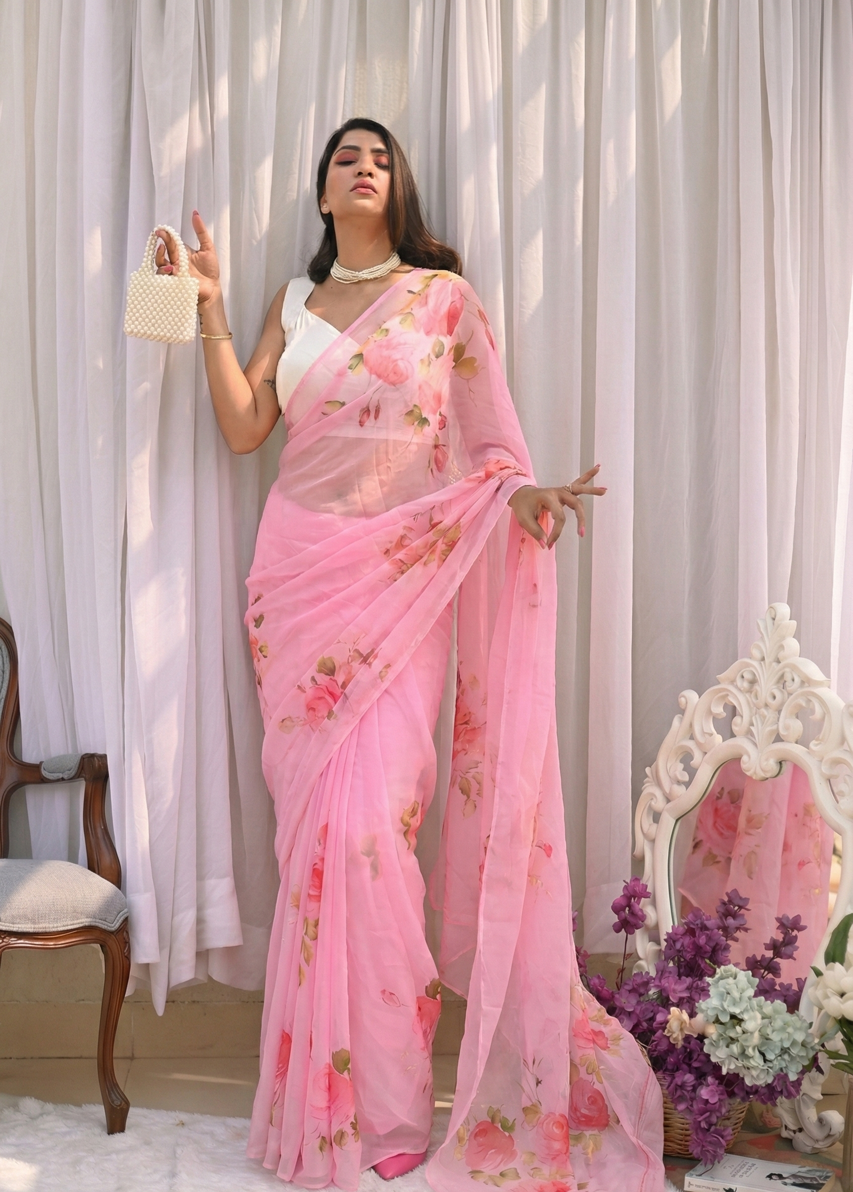 Woman wearing a pink floral saree with a white blouse against a white curtain background.